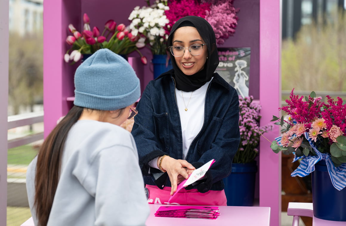 A Random Actor wreathed in pink and red flowers presents an instant photo to a passing park goer
