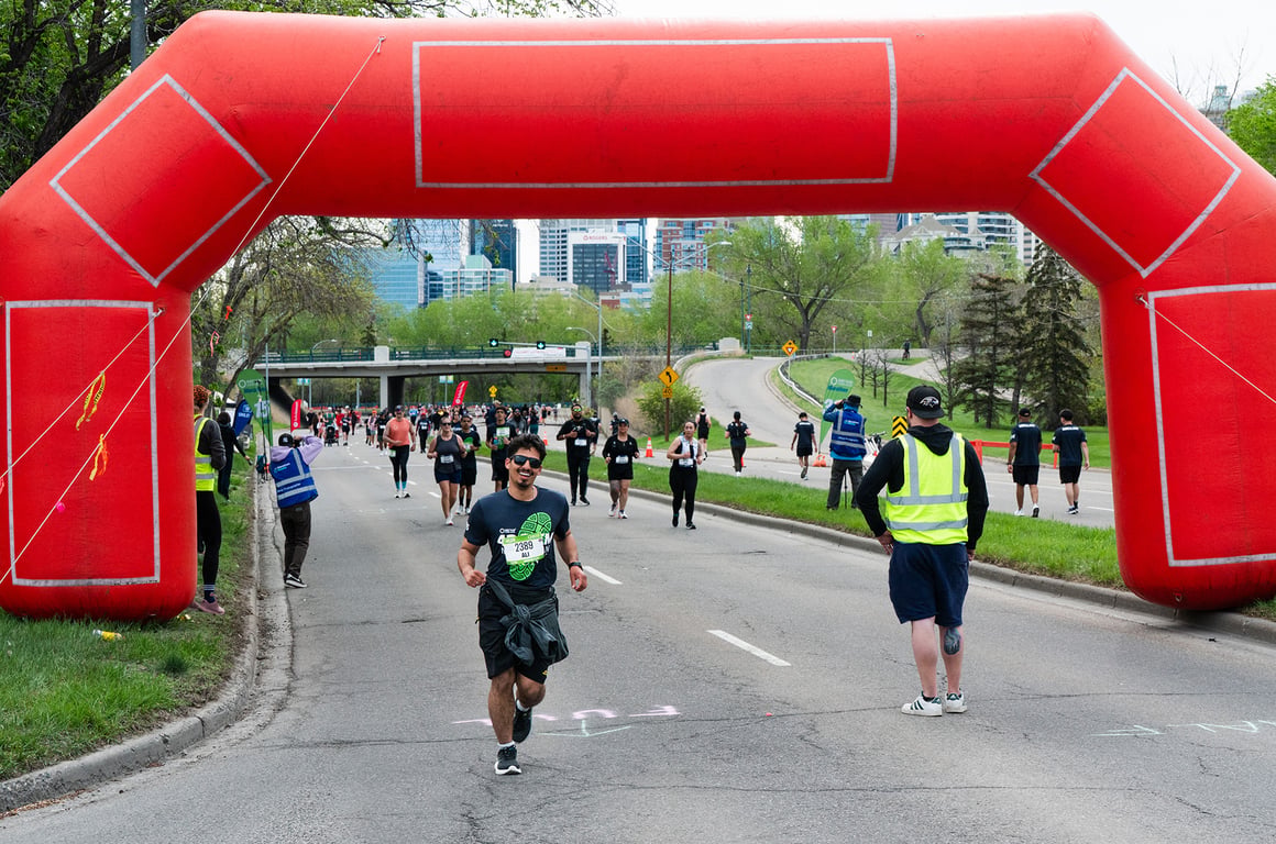 A grinning runner in sunglasses dashes under a big red inflatable arch with other runners in the background.