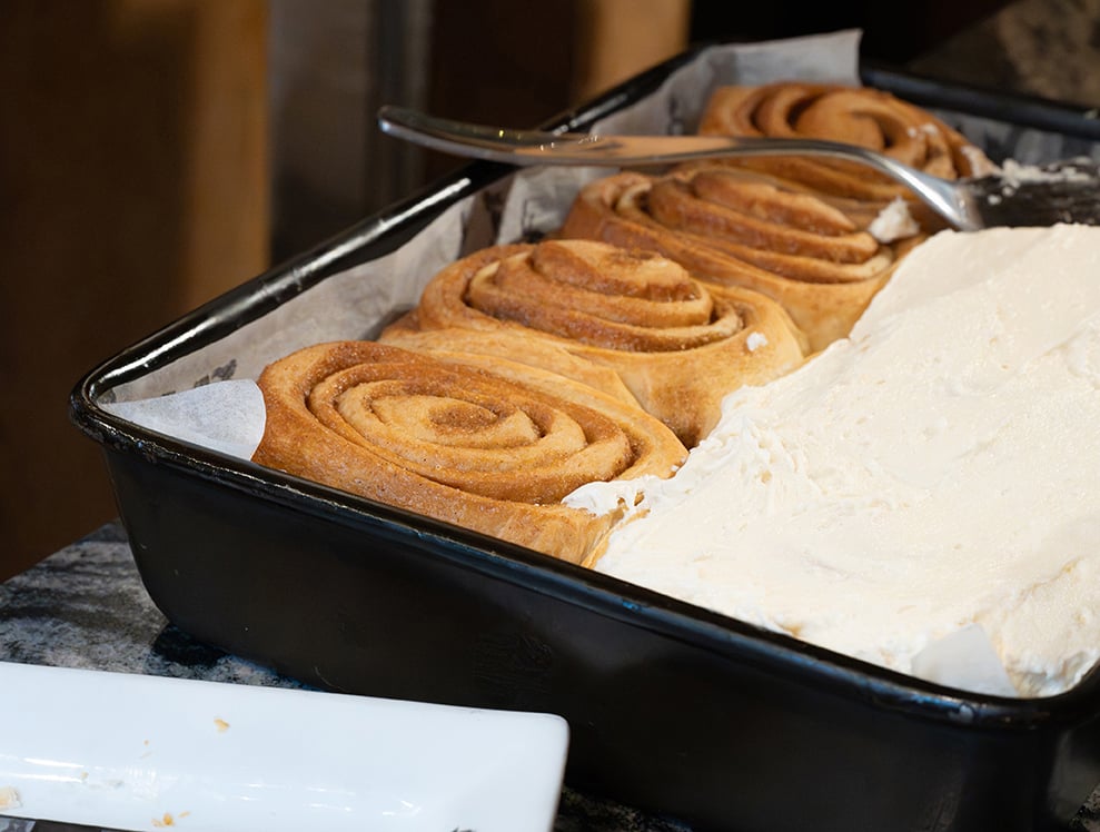A baking tray of frosted, golden cinnamon rolls cools on a marble countertop.